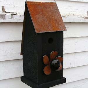 May include: A black wooden birdhouse with a rusty metal roof and a flower-shaped design on the front. The flower is made of rusty metal and has a black glass center.