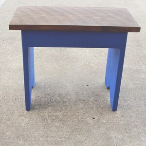 Puede incluir: Un peque&ntilde;o taburete de madera con una parte superior rectangular y cuatro patas. La parte superior es de color madera natural, mientras que las patas y el marco est&aacute;n pintados de azul vibrante. El taburete est&aacute; dise&ntilde;ado para uso pr&aacute;ctico.