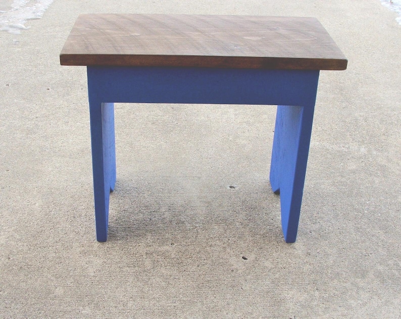 Puede incluir: Un peque&ntilde;o taburete de madera con una parte superior rectangular y cuatro patas. La parte superior es de color madera natural, mientras que las patas y el marco est&aacute;n pintados de azul vibrante. El taburete est&aacute; dise&ntilde;ado para uso pr&aacute;ctico.