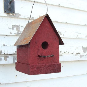 K&ouml;nnte beinhalten: Ein rotes Vogelhaus aus Holz mit einem rostigen Metalldach. Das Vogelhaus hat ein rundes Eingangsloch und h&auml;ngt an einem Draht.