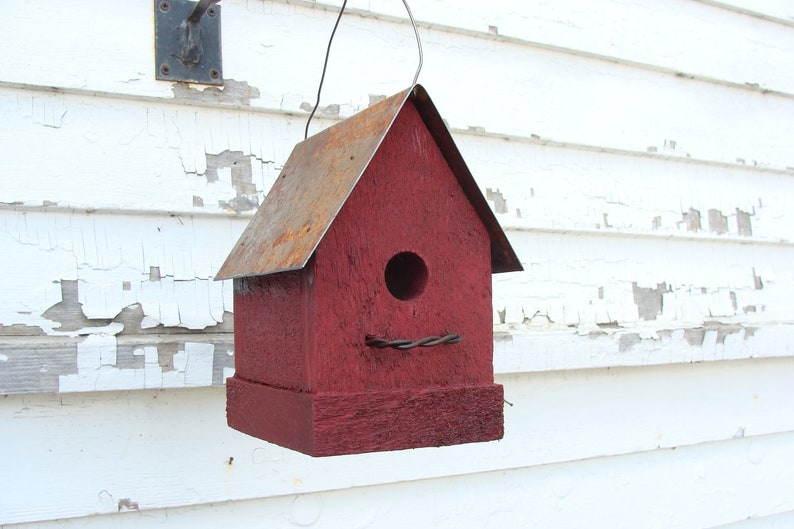 K&ouml;nnte beinhalten: Ein rotes Vogelhaus aus Holz mit einem rostigen Metalldach. Das Vogelhaus hat ein rundes Eingangsloch und h&auml;ngt an einem Draht.
