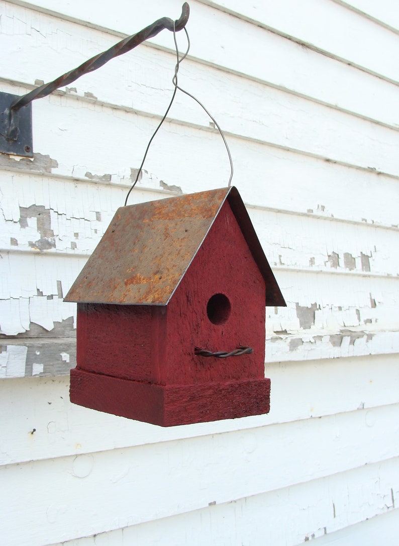 K&ouml;nnte beinhalten: Ein rotes Vogelhaus aus Holz mit einem rostigen Metalldach h&auml;ngt an einem Metallhaken an einer wei&szlig;en Holzwand. Das Vogelhaus hat eine kleine runde &Ouml;ffnung, durch die V&ouml;gel eintreten k&ouml;nnen.