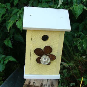 May include: A yellow birdhouse with a white roof and a flower design on the front. The flower is made of metal and has a glass centre.