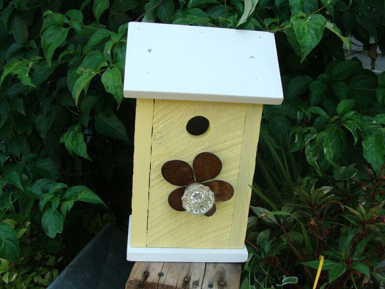 May include: A yellow birdhouse with a white roof and a flower design on the front. The flower is made of metal and has a glass centre.