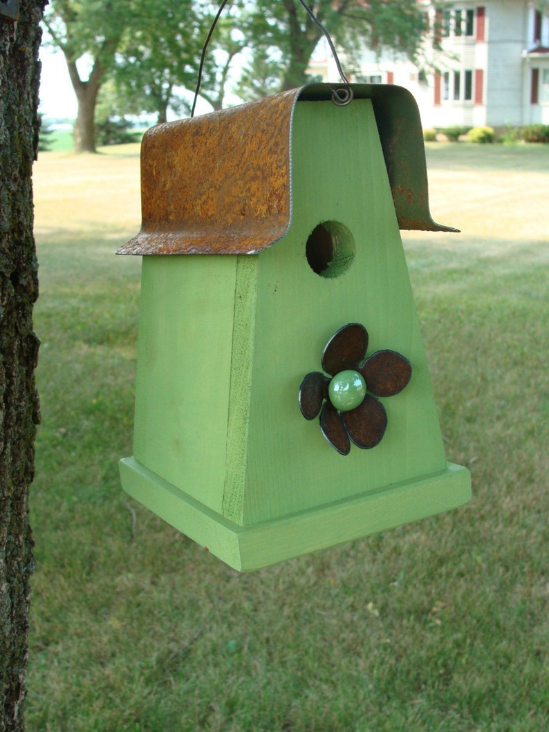 Może przedstawiać: Zielony domek dla ptak&oacute;w z rdzewiejącym metalowym dachem i kwiatowym wzorem z przodu. Domek dla ptak&oacute;w ma mały okrągły otw&oacute;r, przez kt&oacute;ry ptaki mogą wejść.