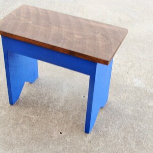 Puede incluir: Un peque&ntilde;o banco de madera con una parte superior rectangular y patas azules. La parte superior est&aacute; te&ntilde;ida de color marr&oacute;n medio y las patas est&aacute;n pintadas de un azul vibrante. El banco es probablemente hecho a mano y adecuado para varios usos.