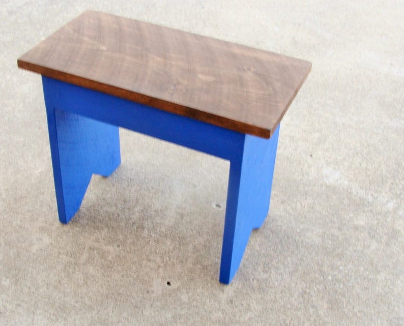 Puede incluir: Un peque&ntilde;o banco de madera con una parte superior rectangular y patas azules. La parte superior est&aacute; te&ntilde;ida de color marr&oacute;n medio y las patas est&aacute;n pintadas de un azul vibrante. El banco es probablemente hecho a mano y adecuado para varios usos.