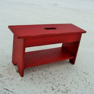 May include: A red wooden bench with a shelf. The bench has a cutout in the top.