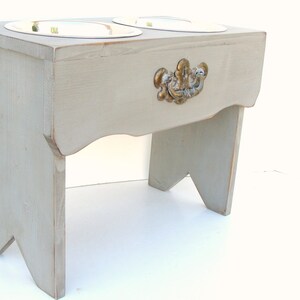 Puede incluir: Un soporte de comida para mascotas de madera gris con dos cuencos de acero inoxidable y una manija dorada decorativa.