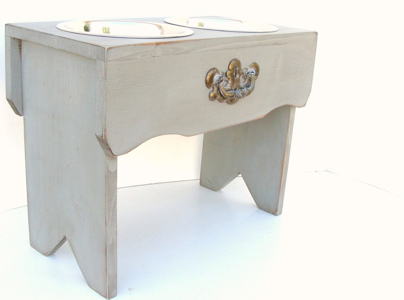 Puede incluir: Un soporte de comida para mascotas de madera gris con dos cuencos de acero inoxidable y una manija dorada decorativa.