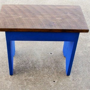 Puede incluir: Un peque&ntilde;o taburete de madera con una parte superior rectangular y cuatro patas. La parte superior est&aacute; te&ntilde;ida de marr&oacute;n oscuro, mientras que las patas est&aacute;n pintadas de azul vibrante. El taburete est&aacute; dise&ntilde;ado para uso en interiores.