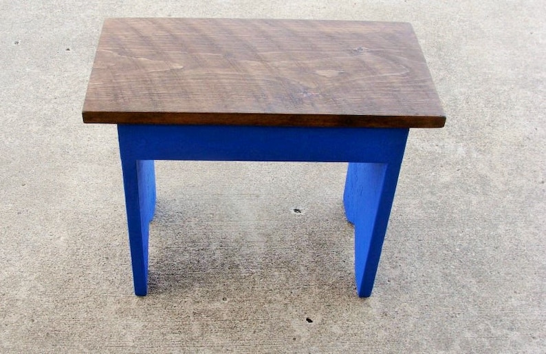 Puede incluir: Un peque&ntilde;o taburete de madera con una parte superior rectangular y cuatro patas. La parte superior est&aacute; te&ntilde;ida de marr&oacute;n oscuro, mientras que las patas est&aacute;n pintadas de azul vibrante. El taburete est&aacute; dise&ntilde;ado para uso en interiores.