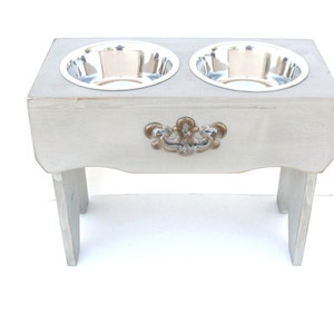 Puede incluir: Un soporte elevado para cuencos de perro de madera gris con dos cuencos de acero inoxidable. El soporte tiene un asa decorativa de color dorado.