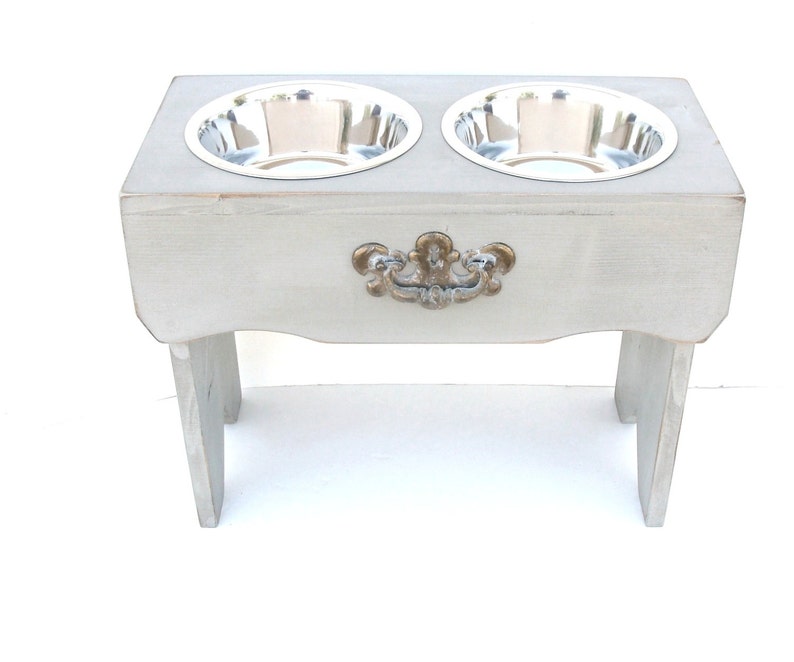 Puede incluir: Un soporte elevado para cuencos de perro de madera gris con dos cuencos de acero inoxidable. El soporte tiene un asa decorativa de color dorado.