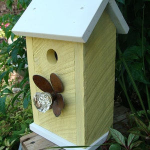 May include: A yellow birdhouse with a white roof and white trim. The birdhouse has a small hole for birds to enter and a decorative glass knob with a rusty metal design.
