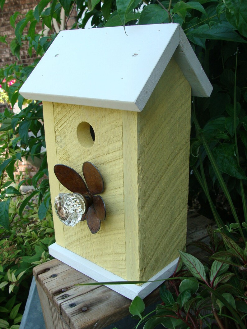 May include: A yellow birdhouse with a white roof and white trim. The birdhouse has a small hole for birds to enter and a decorative glass knob with a rusty metal design.