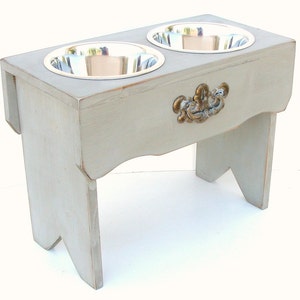 Puede incluir: Un soporte elevado para cuencos de perro de madera gris con dos cuencos de acero inoxidable y una manija decorativa de metal.
