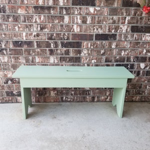 May include: A light green wooden bench with a simple design. The bench has a rectangular seat and two legs on each side. The bench is made of solid wood and has a natural finish.