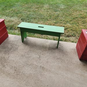 May include: A green wooden bench with a small hole in the top, sitting on a concrete patio. Two red wooden planters are on either side of the bench.