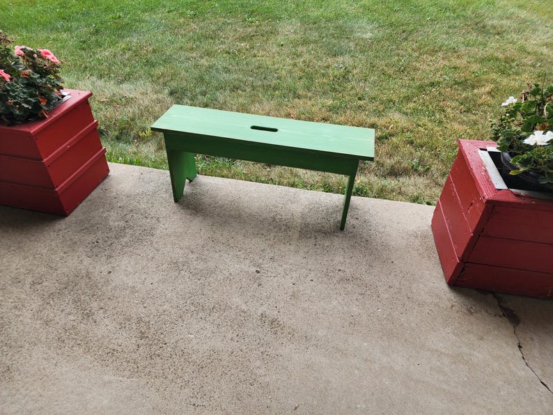 May include: A green wooden bench with a small hole in the top, sitting on a concrete patio. Two red wooden planters are on either side of the bench.