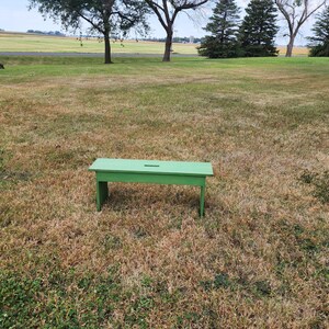May include: A green wooden bench with a small cutout in the top. The bench is sitting on a grassy field.