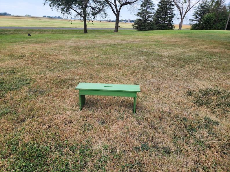 May include: A green wooden bench with a small cutout in the top. The bench is sitting on a grassy field.