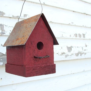 K&ouml;nnte beinhalten: Ein rotes Vogelhaus aus Holz mit einem rostigen Metalldach. Das Vogelhaus hat ein rundes Loch, durch das die V&ouml;gel eintreten k&ouml;nnen, und eine Drahtritze.
