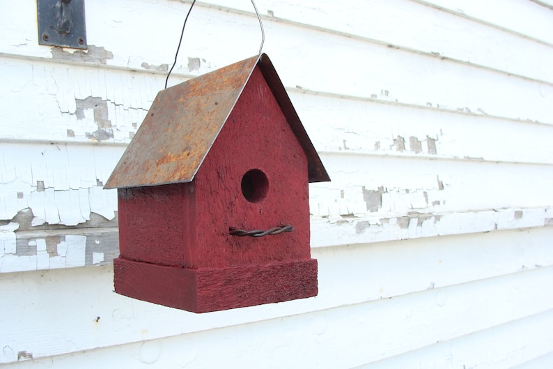 K&ouml;nnte beinhalten: Ein rotes Vogelhaus aus Holz mit einem rostigen Metalldach. Das Vogelhaus hat ein rundes Loch, durch das die V&ouml;gel eintreten k&ouml;nnen, und eine Drahtritze.