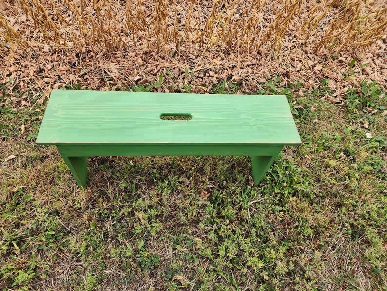 May include: A green wooden bench with a cutout in the top. The bench is sitting on a bed of green grass.