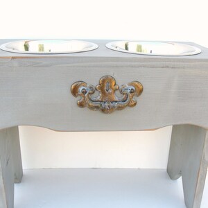 Puede incluir: Un soporte elevado de madera gris para cuencos de comida y agua para mascotas con dos cuencos de acero inoxidable. El soporte tiene una manija decorativa dorada.