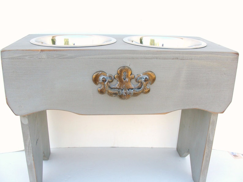 Puede incluir: Un soporte elevado de madera gris para cuencos de comida y agua para mascotas con dos cuencos de acero inoxidable. El soporte tiene una manija decorativa dorada.