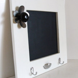 May include: White framed chalkboard with a black writing surface. Features a decorative black flower accent and two silver hooks below. A small silver dish is also present. Ideal for home decor.