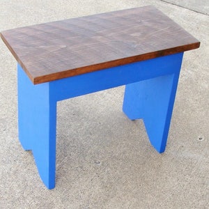 Puede incluir: Un peque&ntilde;o taburete de madera con una parte superior rectangular y cuatro patas. La parte superior est&aacute; te&ntilde;ida de marr&oacute;n oscuro, mientras que las patas y el marco est&aacute;n pintados de azul brillante. El taburete mide aproximadamente 30 cm de largo, 20 cm de ancho y 25 cm de alto.