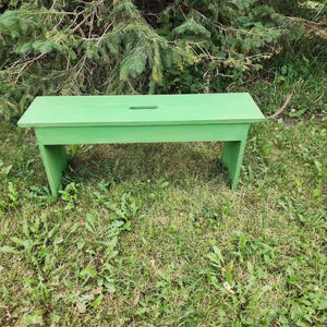 May include: A solid green wooden bench with a simple design. The bench is made of rough-hewn wood and has a natural finish. It is sitting on a bed of green grass.