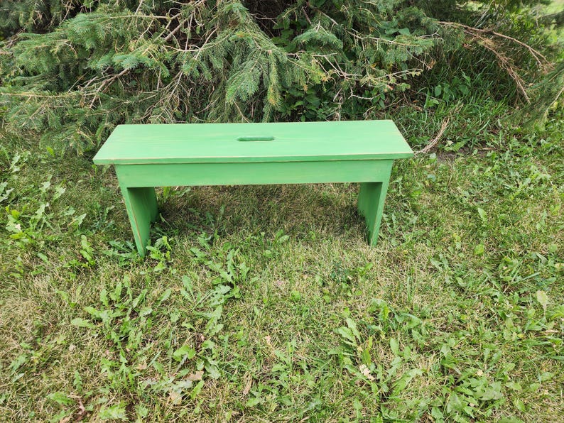 May include: A solid green wooden bench with a simple design. The bench is made of rough-hewn wood and has a natural finish. It is sitting on a bed of green grass.