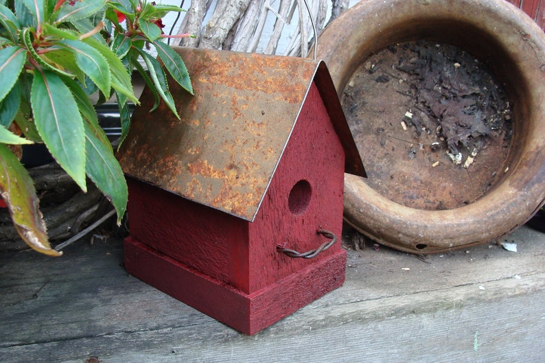 K&ouml;nnte beinhalten: Ein rotes Vogelhaus aus Holz mit einem rostigen Metalldach. Das Vogelhaus hat eine kleine &Ouml;ffnung f&uuml;r V&ouml;gel, um einzudringen, und eine Sitzstange vorne.