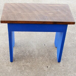 Puede incluir: Un peque&ntilde;o taburete de madera con una parte superior rectangular y cuatro patas. La parte superior est&aacute; te&ntilde;ida de marr&oacute;n oscuro, mientras que las patas y el marco est&aacute;n pintados de azul vibrante. El taburete est&aacute; dise&ntilde;ado para uso en interiores o exteriores.