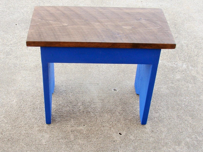 Puede incluir: Un peque&ntilde;o taburete de madera con una parte superior rectangular y cuatro patas. La parte superior est&aacute; te&ntilde;ida de marr&oacute;n oscuro, mientras que las patas y el marco est&aacute;n pintados de azul vibrante. El taburete est&aacute; dise&ntilde;ado para uso en interiores o exteriores.