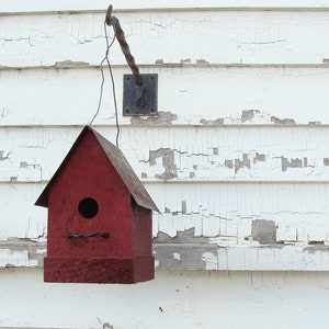 K&ouml;nnte beinhalten: Ein rotes Vogelhaus mit einer kleinen &Ouml;ffnung f&uuml;r V&ouml;gel, um hineinzugelangen. Das Vogelhaus h&auml;ngt an einem Metallhaken, der an einer wei&szlig;en Holzwand mit abbl&auml;tternder Farbe befestigt ist.