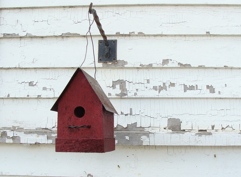 K&ouml;nnte beinhalten: Ein rotes Vogelhaus mit einer kleinen &Ouml;ffnung f&uuml;r V&ouml;gel, um hineinzugelangen. Das Vogelhaus h&auml;ngt an einem Metallhaken, der an einer wei&szlig;en Holzwand mit abbl&auml;tternder Farbe befestigt ist.