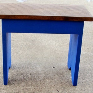 Puede incluir: Un peque&ntilde;o taburete de madera con una parte superior rectangular y cuatro patas. La parte superior es de color marr&oacute;n oscuro, mientras que las patas y el marco est&aacute;n pintados de azul vibrante. El taburete mide aproximadamente 30 cm de largo, 25 cm de ancho y 25 cm de alto.