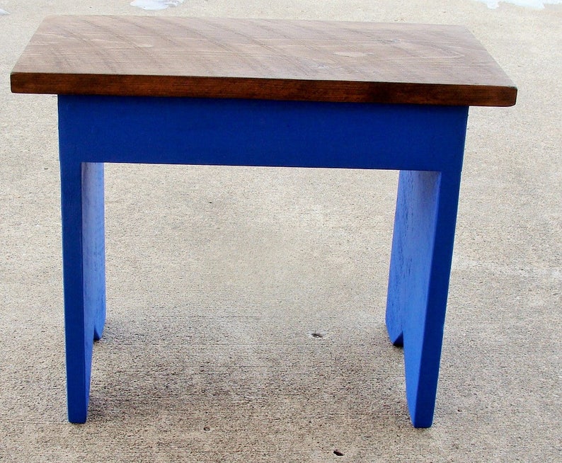 Puede incluir: Un peque&ntilde;o taburete de madera con una parte superior rectangular y cuatro patas. La parte superior es de color marr&oacute;n oscuro, mientras que las patas y el marco est&aacute;n pintados de azul vibrante. El taburete mide aproximadamente 30 cm de largo, 25 cm de ancho y 25 cm de alto.