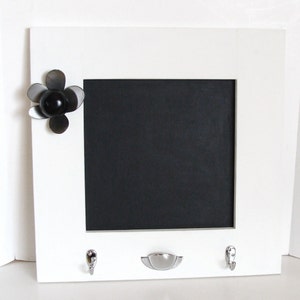 May include: A white framed chalkboard with a black center, featuring a decorative metal flower and two silver hooks. A silver drawer pull is below the chalkboard. This item is designed for home organization and decor.