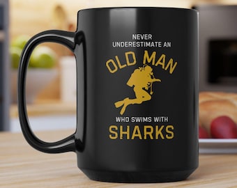 Puede incluir: Taza de café de cerámica negra con un asa grande. La taza presenta el texto "NEVER UNDERESTIMATE AN OLD MAN WHO SWIMS WITH SHARKS" en letras doradas, junto con una silueta de buzo. La taza está sobre una superficie de madera.