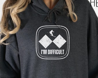 Puede incluir: Sudadera con capucha gris oscuro jaspeado con un gráfico blanco. El gráfico presenta un esquiador, dos rombos y el texto "I'M DIFFICULT". La sudadera tiene una capucha con cordón y un bolsillo delantero.