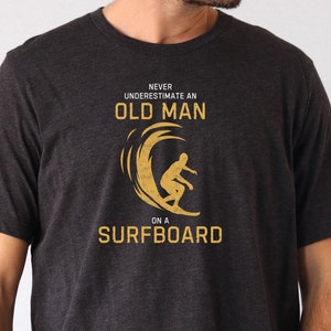 May include: Dark grey heather t-shirt with a graphic of a surfer riding a wave and the text "Never underestimate an old man on a surfboard".