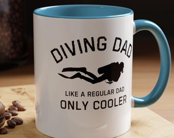 Puede incluir: Taza de cerámica blanca con interior y asa azules. La taza presenta el texto "DIVING DAD" sobre una silueta de buceador, con la frase "LIKE A REGULAR DAD ONLY COOLER" debajo. Una cuchara y un macaron están en primer plano.