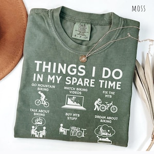 May include: Moss green t-shirt with white text and illustrations about mountain biking. The shirt reads "THINGS I DO IN MY SPARE TIME" with related activities. A gold necklace with a pendant rests on the shirt.
