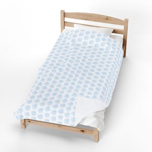 May include: A light wooden bed frame featuring a white duvet cover with a repeating pattern of small, light blue seashell illustrations. The bedding is neatly tucked and draped over the mattress, showcasing a clean, simple design on the fabric.