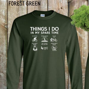 May include: A forest green long-sleeved t-shirt with a white graphic that says "Things I do in my spare time" and lists activities related to biking.